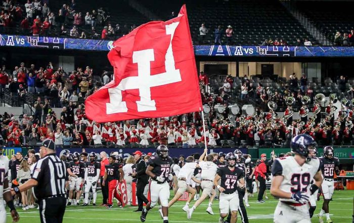 Two high school football players jog across a field. In the background, two boys run by with a flag including the Lake Highlands High School logo.