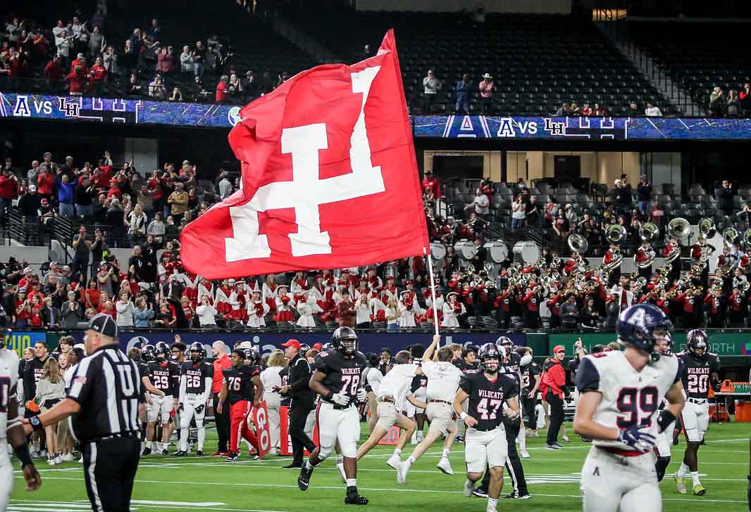 Two high school football players jog across a field. In the background, two boys run by with a flag including the Lake Highlands High School logo.