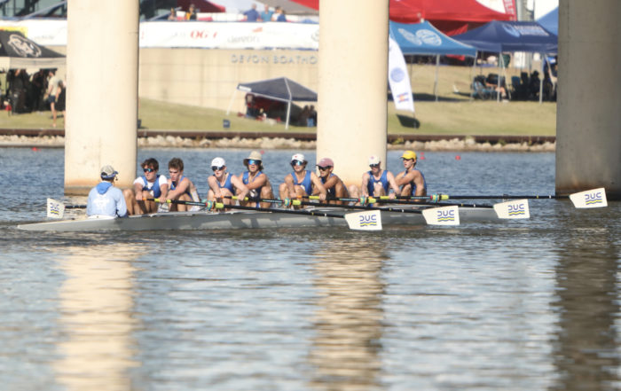 Dallas United Crew varsity boys rowing. There are eight of them, and the blades of their oars are in the air as they prepare to take a stroke.