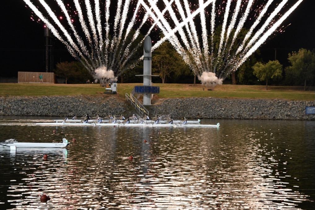 A boat of eight rowers and one coxswain crosses a finish line with two boats right behind them. Fireworks go of in the background. It is night.