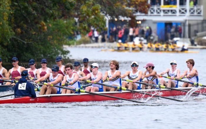 a rowing boat of eight rowers and one coxswain rows past a rival boat. they are preparing to turn a corner on the river. another crew is ahead of them.