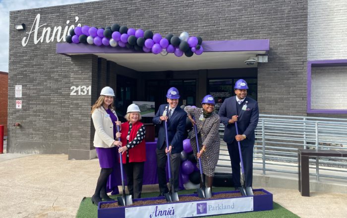 From left to right, two women, one man, another woman, and a man dig into a rectangular planter of dirt in front of a grey and purple building. A sign says "Annie's Place."
