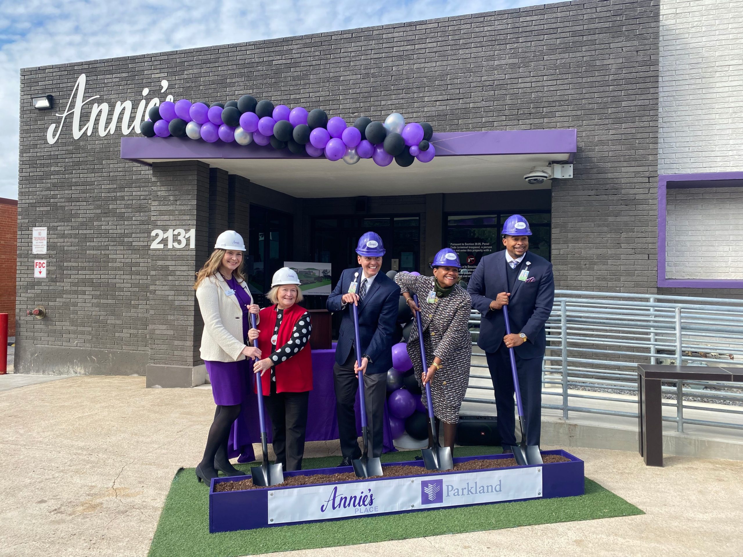 From left to right, two women, one man, another woman, and a man dig into a rectangular planter of dirt in front of a grey and purple building. A sign says "Annie's Place."