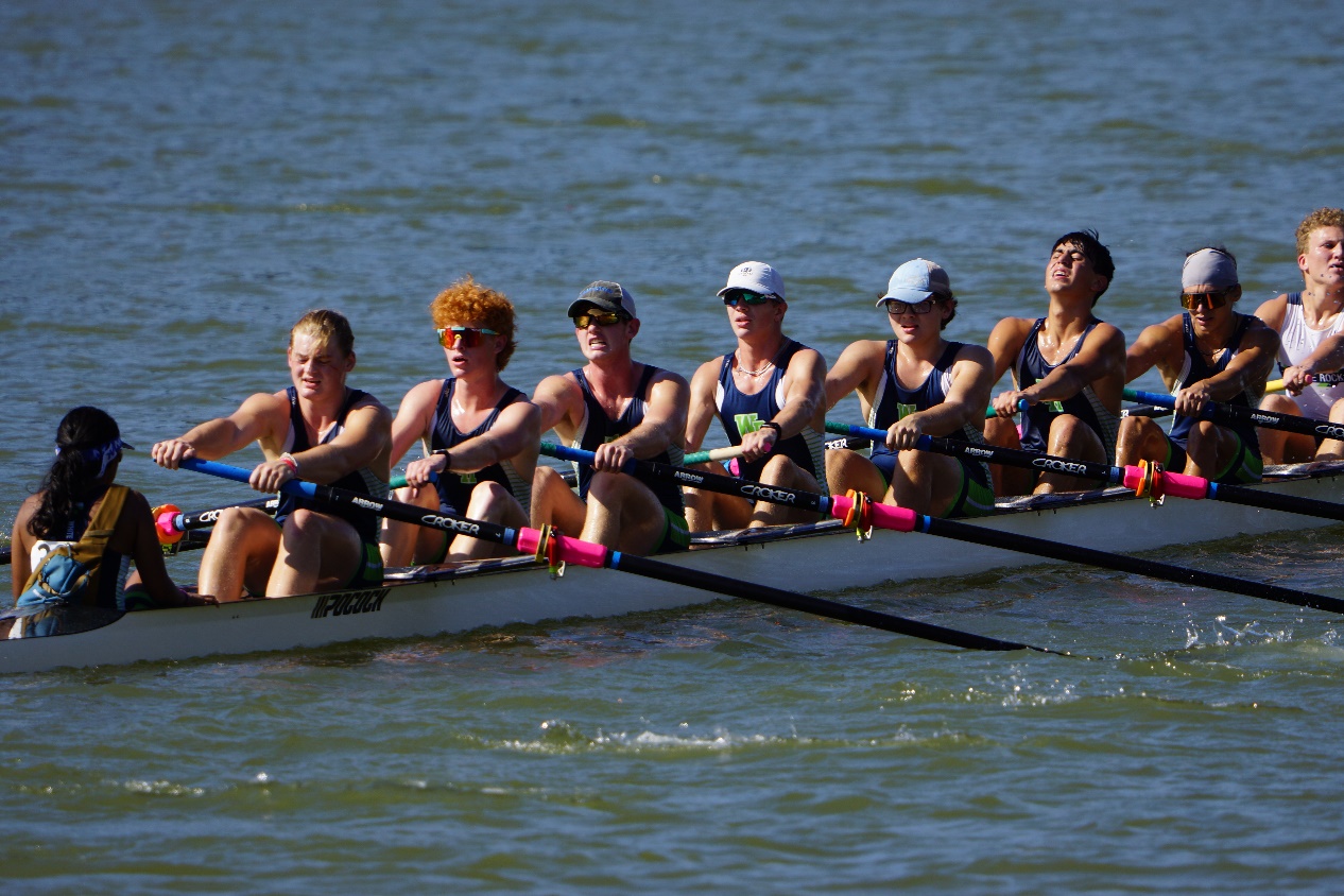 A boat of eight male rowers and one female coxswain rowing at the Waco Rowing Regatta. All of the rowers are grimacing with exertion.